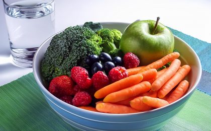 Eine Schale mit frischem Obst und Gemüse, darunter Äpfel, Beeren, Karotten und Brokkoli, ergänzt durch ein Glas Wasser.
