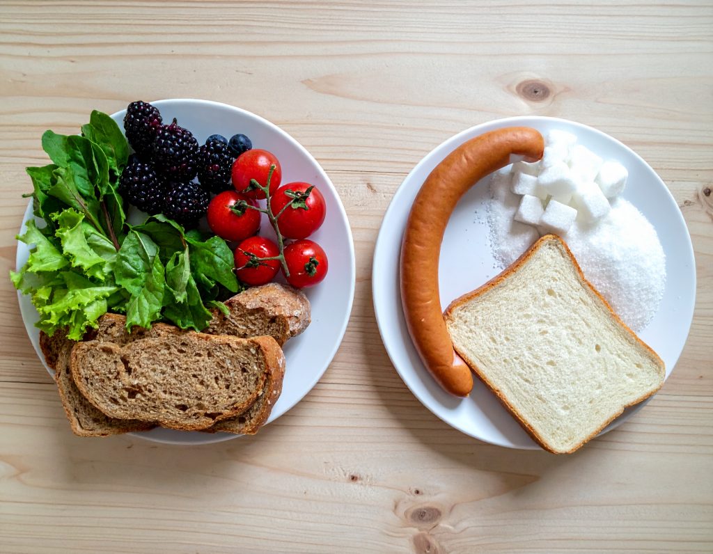 Eine Gegenüberstellung von zwei Tellern: Links Fast Food (Zucker, weißes Brot, Wurst), rechts frische Lebensmittel wie Beeren, Vollkornbrot und grünes Gemüse.