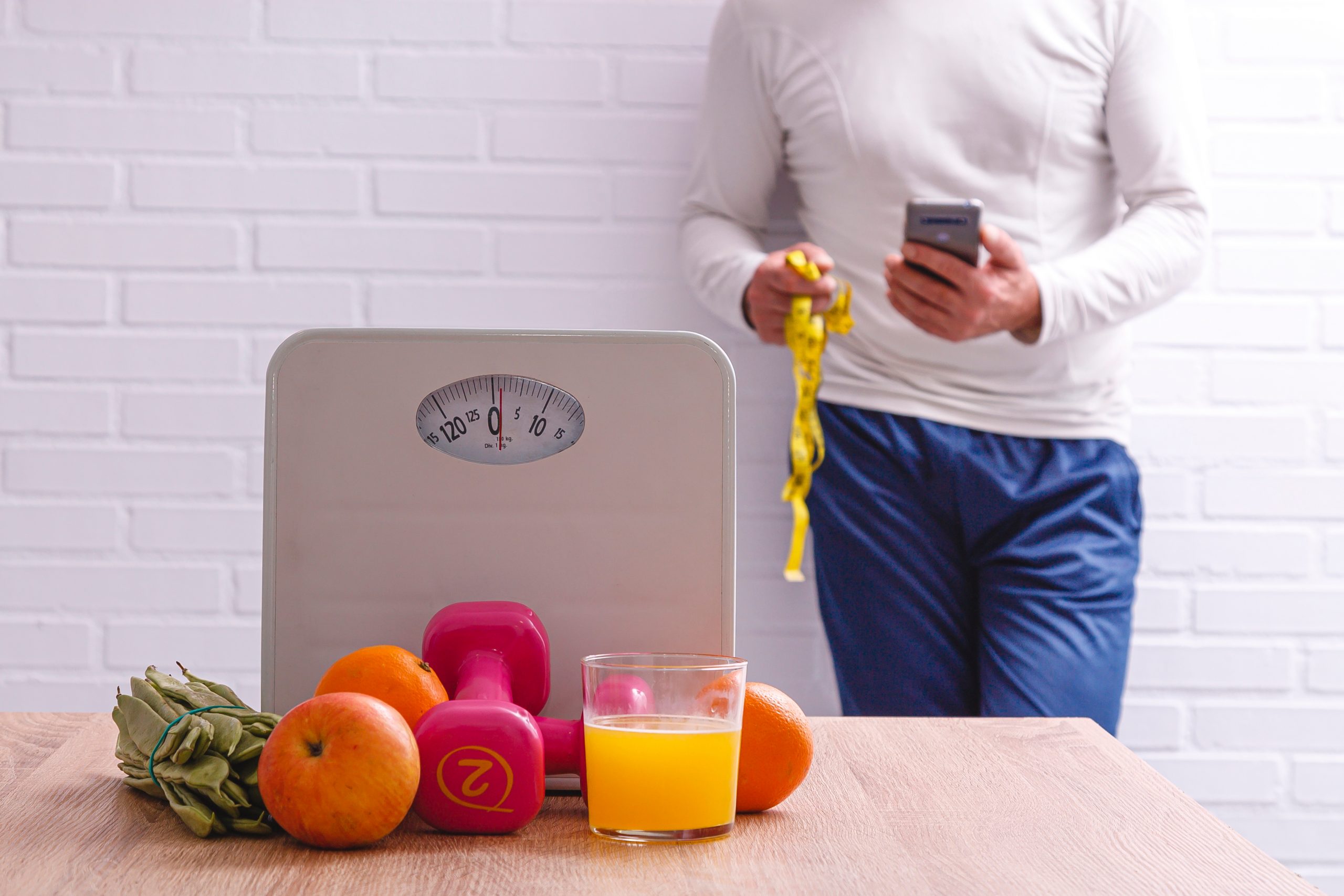 Mann steht hinter einer Waage mit Obst, Hanteln und Orangensaft und überprüft sein Gewicht nach dem Absetzen von Abnehmspritzen.